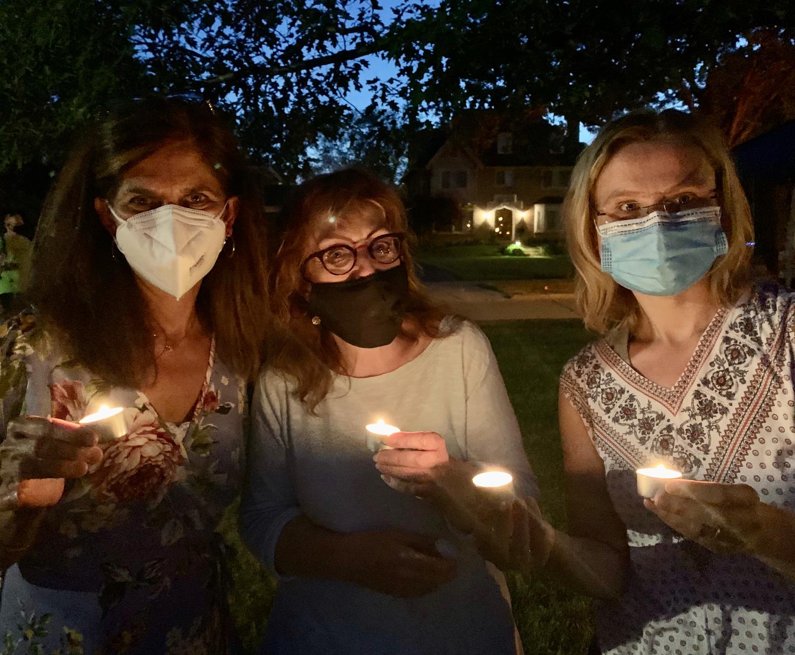 Three women hold candles at dusk. They wear face masks.