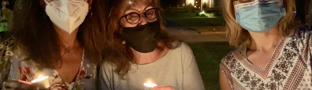 Three women hold candles at dusk. They wear face masks.