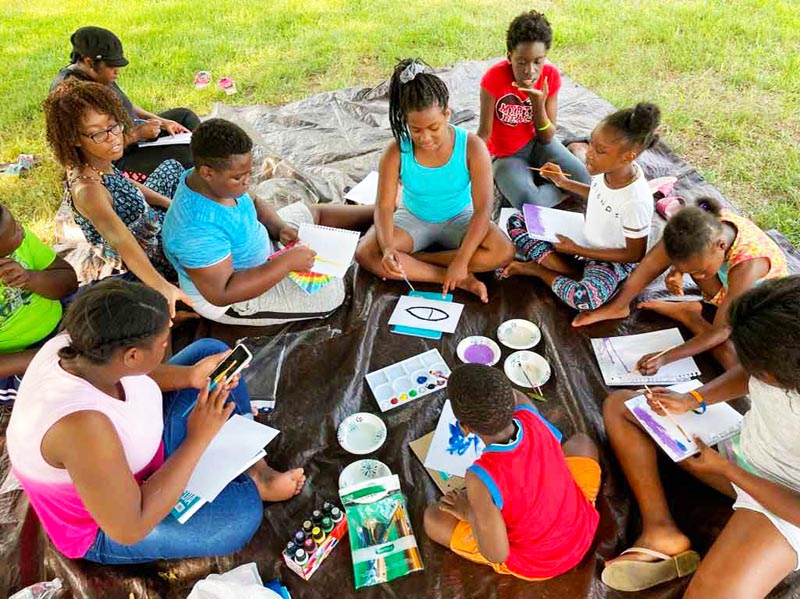 A group of children sits on a blanket outdoors and paints pictures.