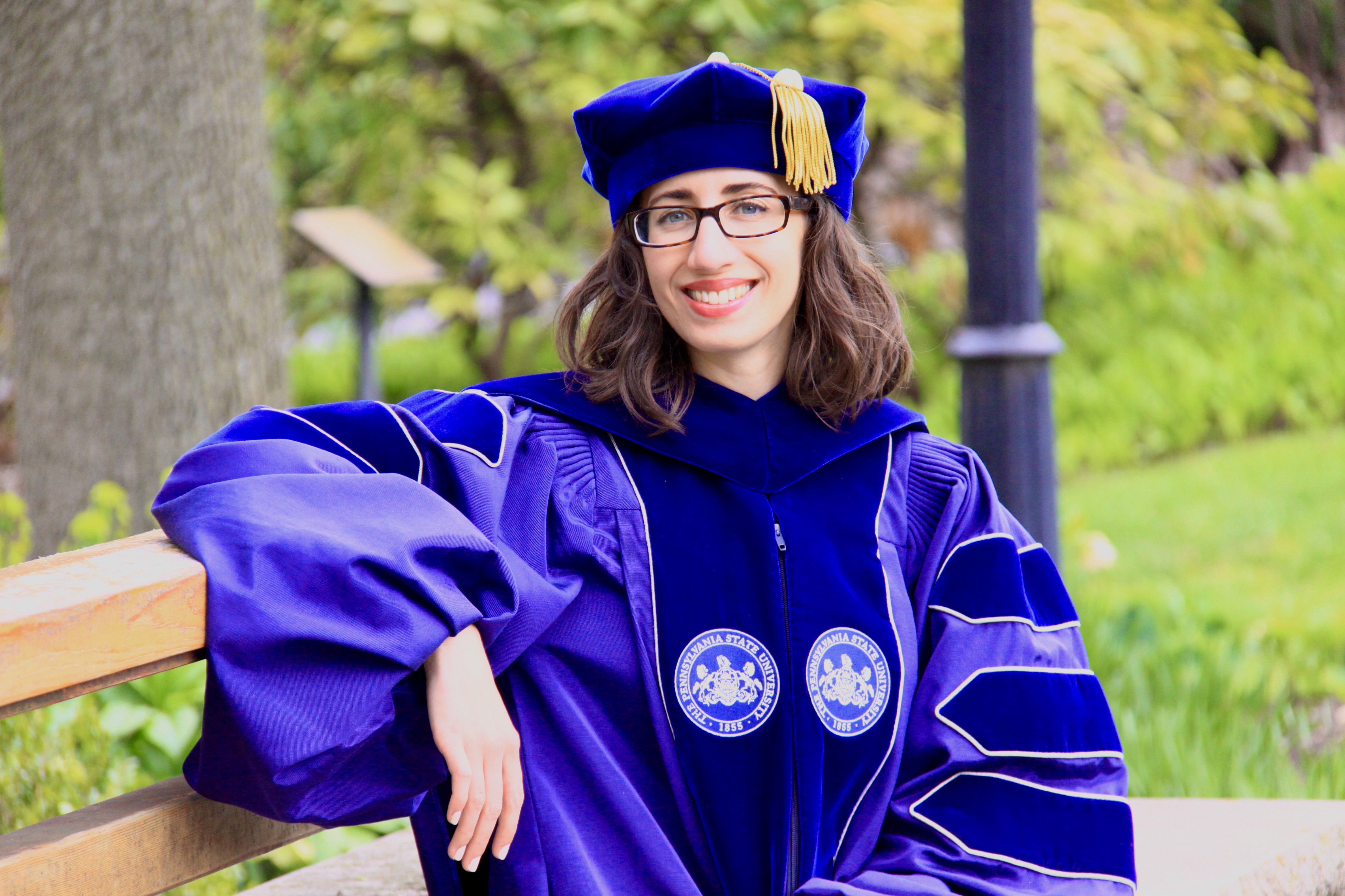 A young woman sits on a bench. She wears academic robes.
