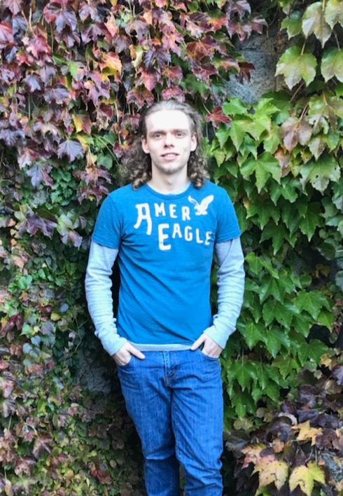 A young man stands in front of foliage and smiles.
