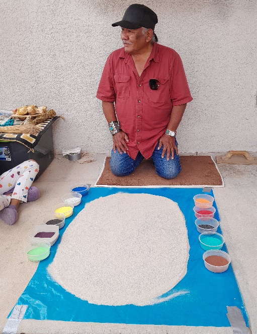 An Indigenous artist kneels in front of a work area with colored sands laid on the ground.