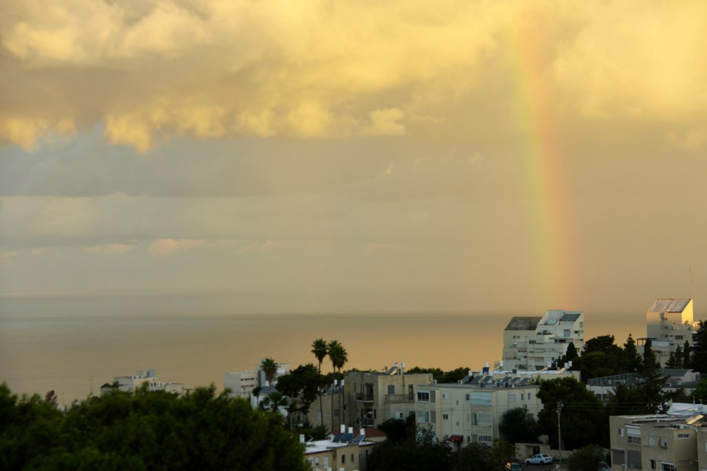 A rainbow arcs over a sea. In the foreground are concrete apartment buildings and foliage.