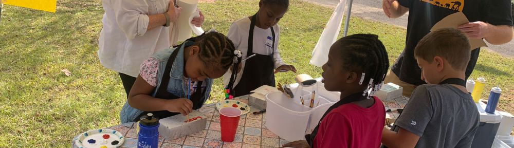 Several children paint messages on bricks with the guidance of two adults.