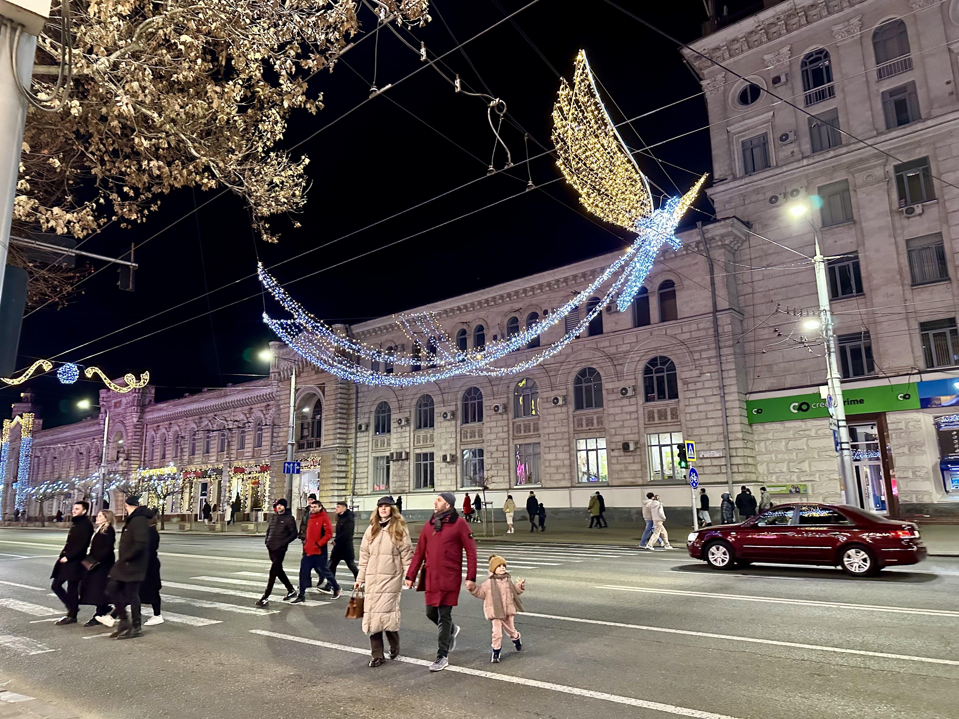 Christmas lights depict an angelic figure, which hangs above historical buildings, a city street, and warmly dressed pedestrians.