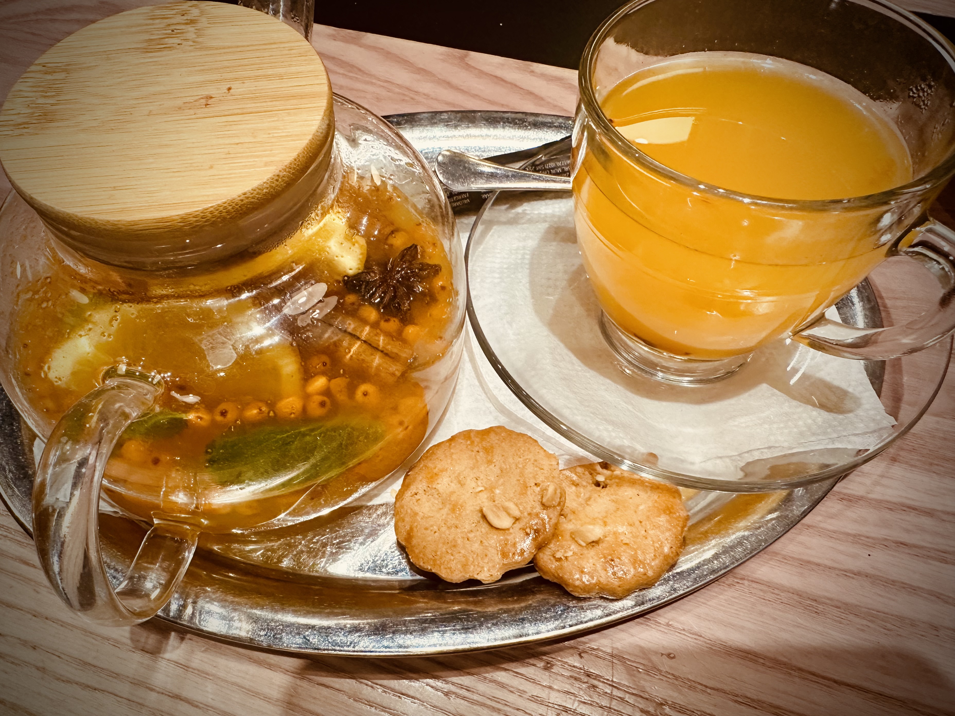 A glass teacup and teapot are filled with an orange beverage. They sit on a silver serving tray with cookies.