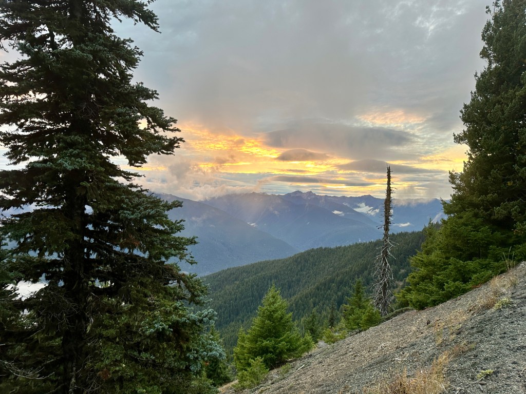 The sun sets behind mountains as seen from a peak.