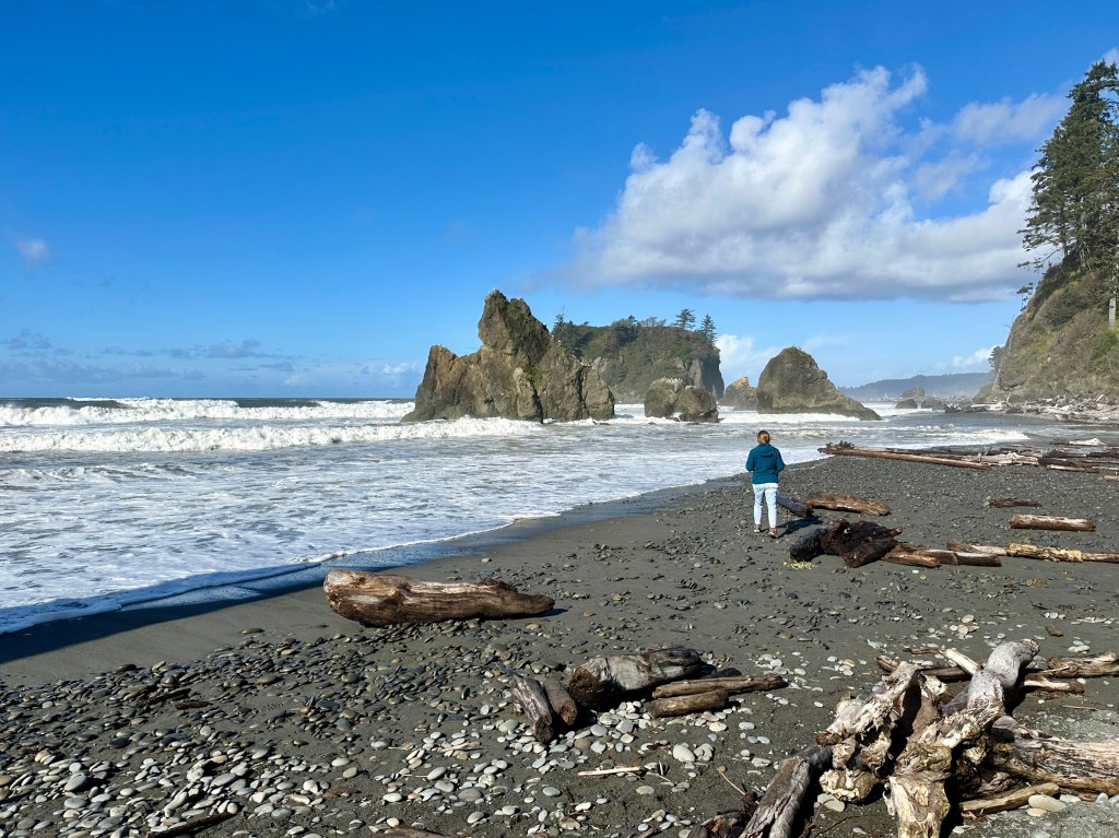 A woman stands on a seashore. Behind her, a tall rock formation rises from the surf.