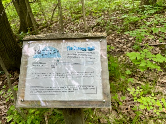 A sign in a forest titled "The Oneness Walk." The sign describes developments lost to history, both spiritual and geological, due to lack of records.